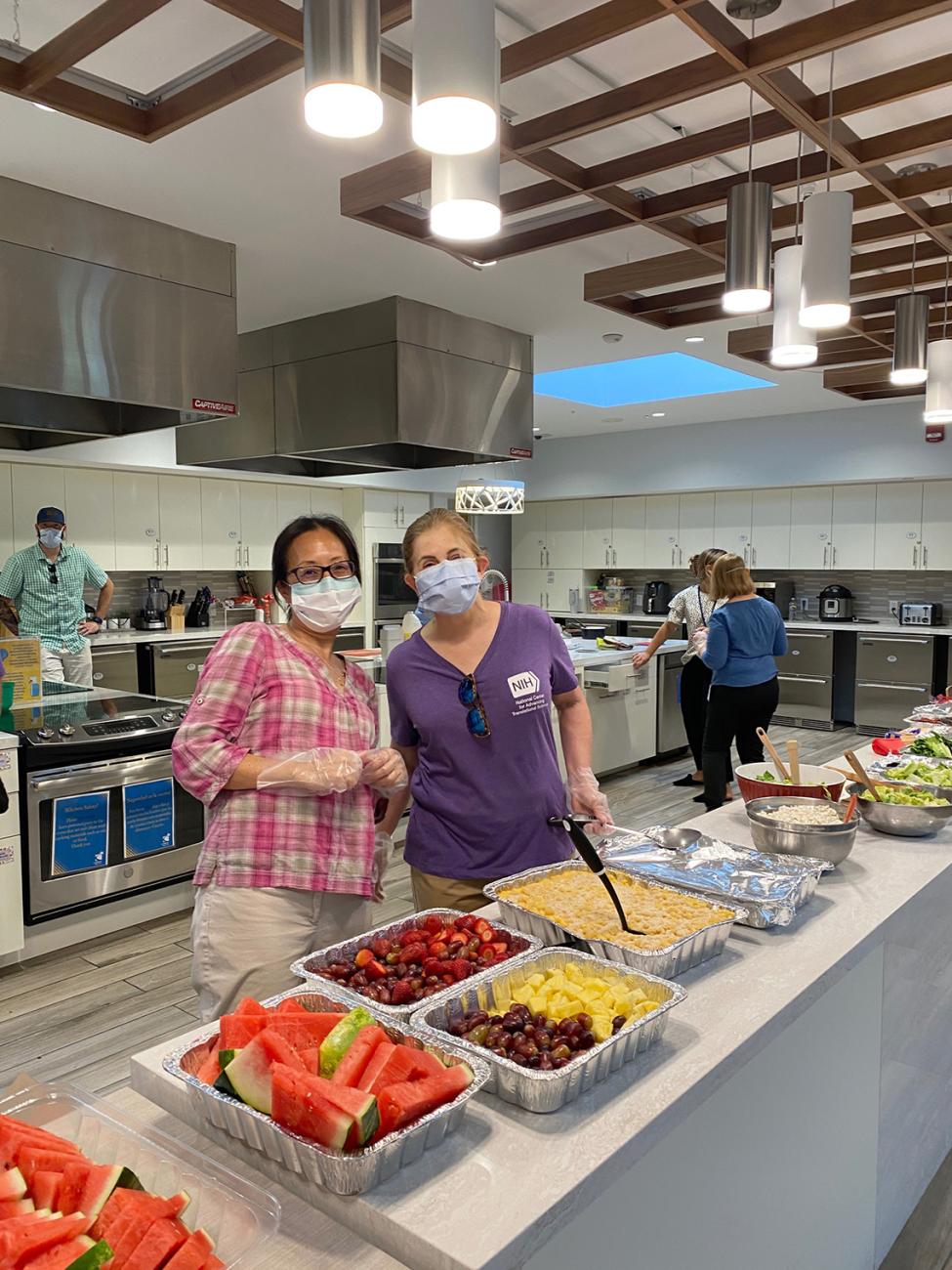 Two masked women stand at a counter ready to serve platters of food