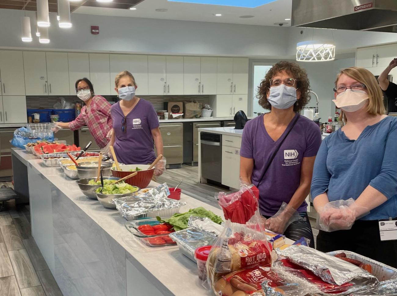 Four masked women stand along a counter lined with food