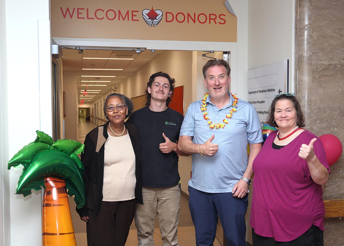Four people smile at the entrance to the new Blood Bank location. Two men give thumbs up gesture.