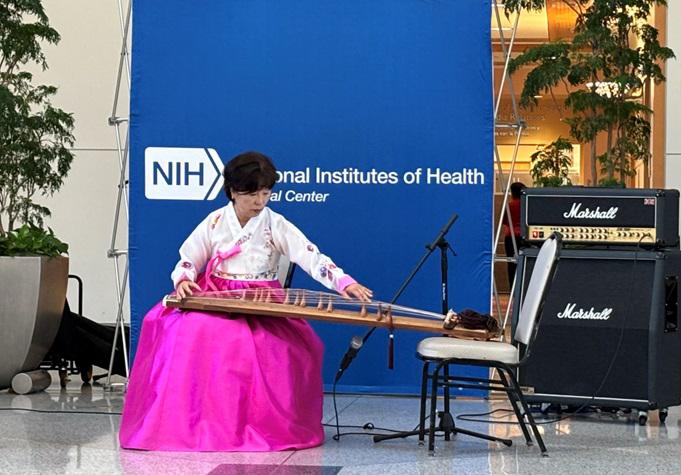 A woman in a white, flowered top and long pink skirt plays a long, wooden string instrument. It is balanced between her lap and a chair.