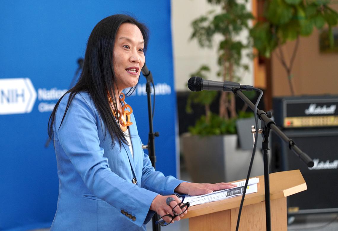 Dark-haired woman speaks at a podium. She is wearing a light blue sweater and orange necklace.