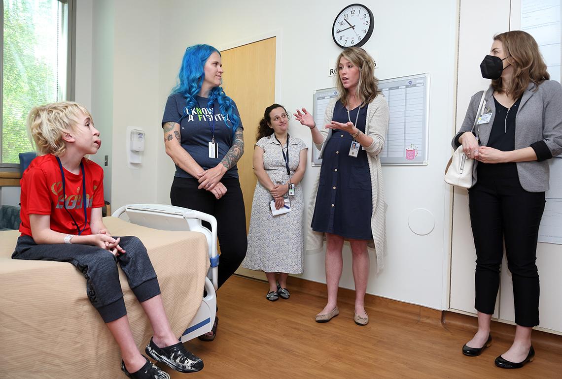 Youngster sits on bed, alongside four adult women. One woman is gesturing with her hands, speaking. One woman is masked, observing.