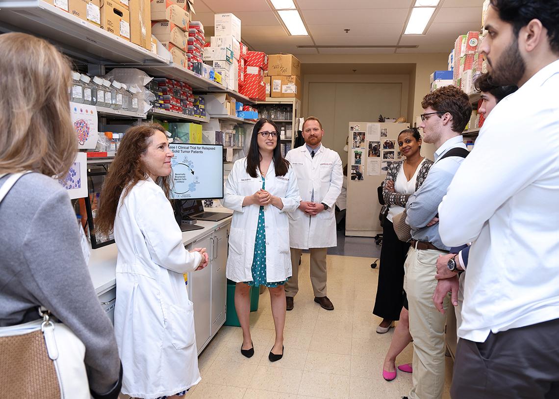 Several people gathered around a woman speaking in a lab.