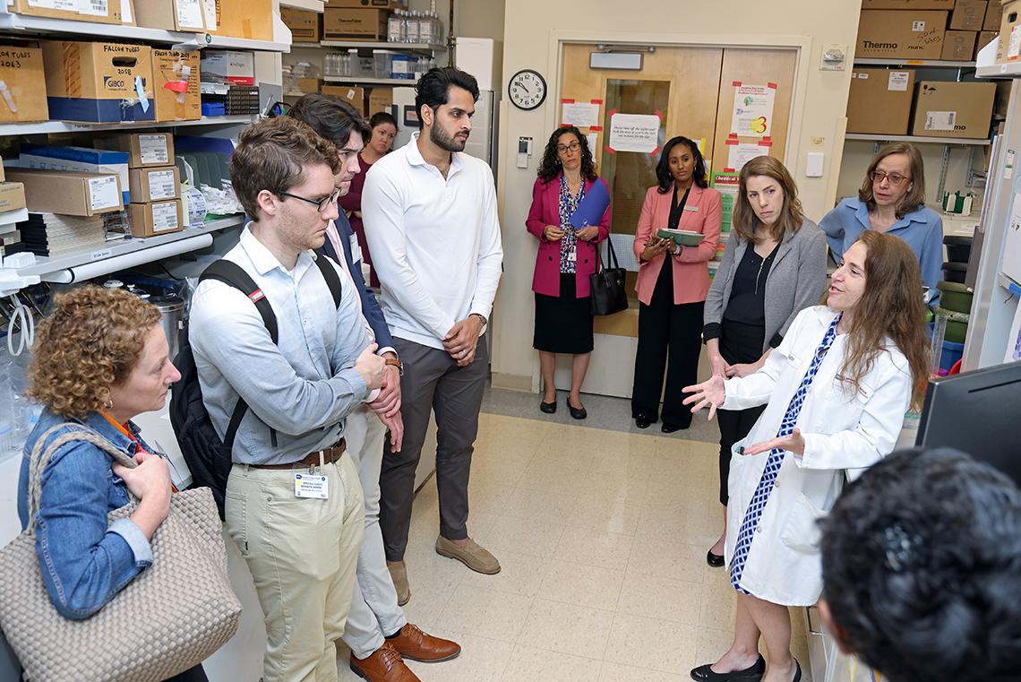 Group of men and women stand in a lab as a woman in a labcoat speaks to them.