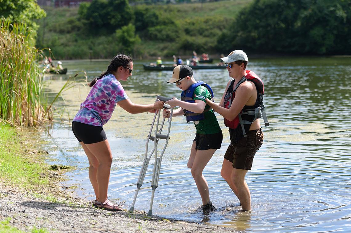 A boy on crutches wades out of a lake with help from two counselors. 