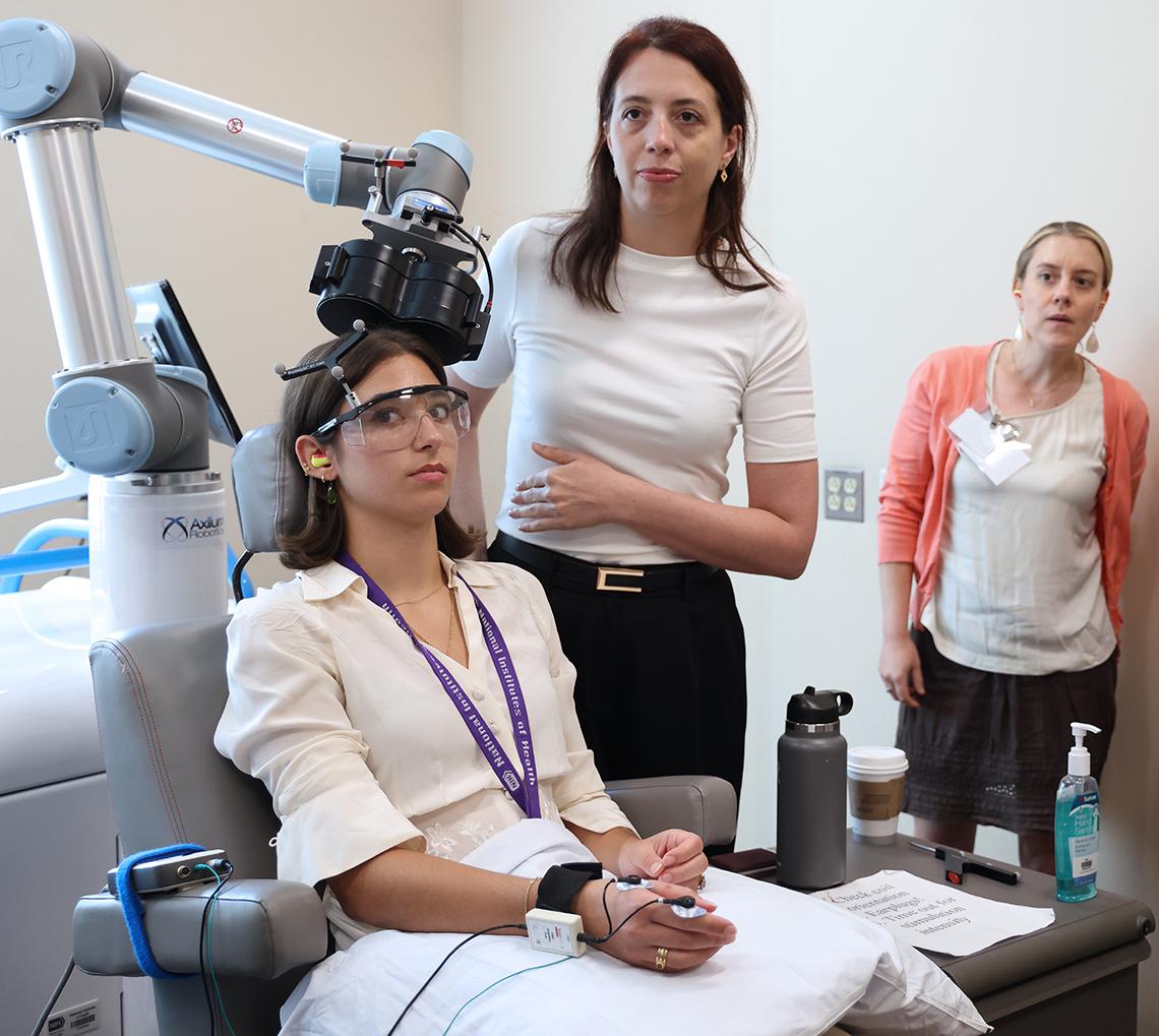 Woman sits under TMS machine with NIH staff standing nearby to assist