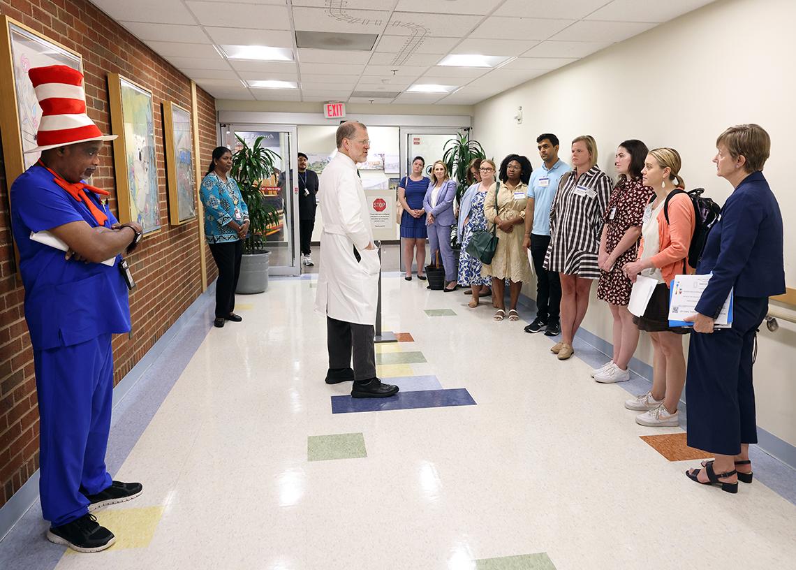 Dr. Pine in lab coat stands in hallway outside CC pediatric unit talking to group lined up by wall, an aid wearing Dr. Seuss red and white striped hat stands opposite.