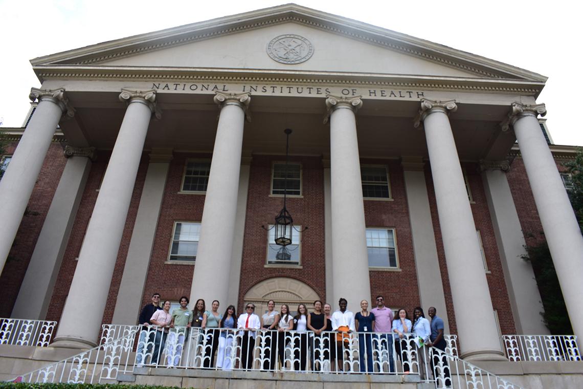 A group of students stand at the forefront of a large brick building with rows of columns.