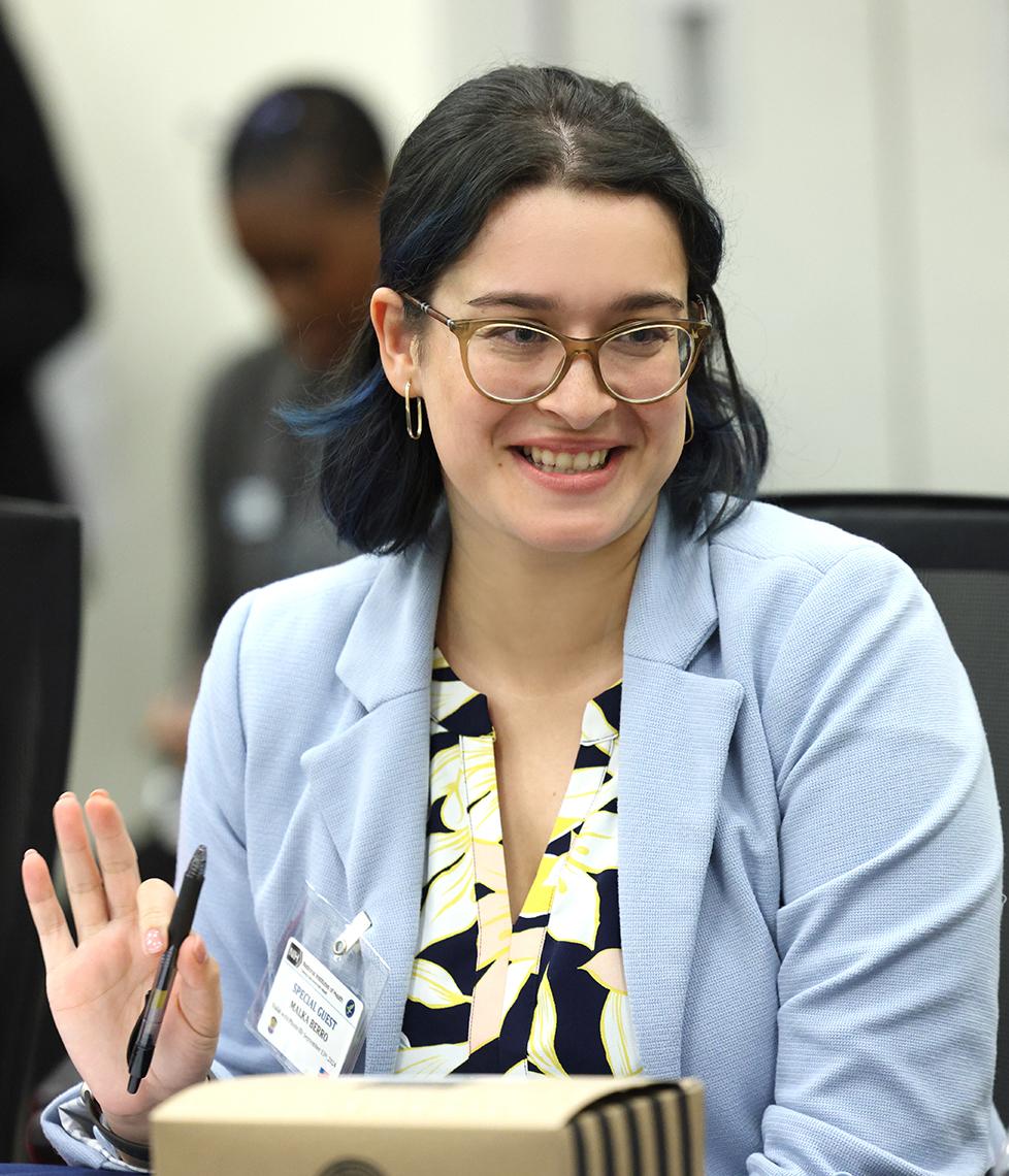 A woman in light blue sweater, wearing glasses, talks.