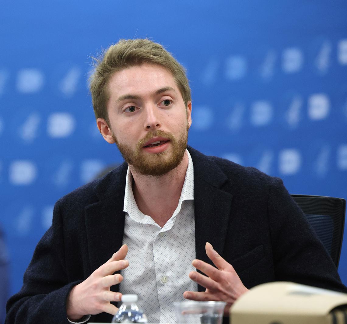 Bearded man talking, with hands extended, against blue NIH backdrop
