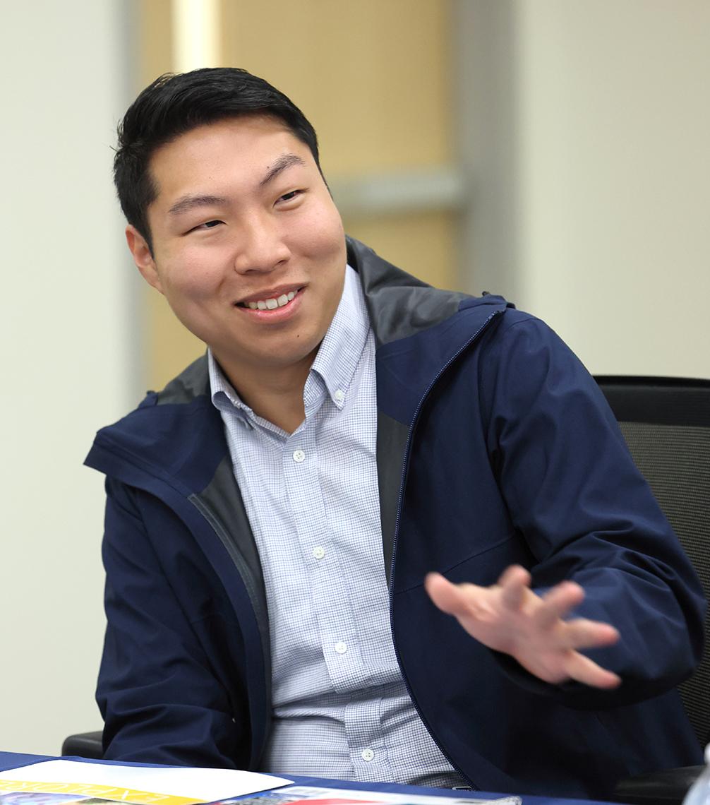 A seated man talks with hand extended.