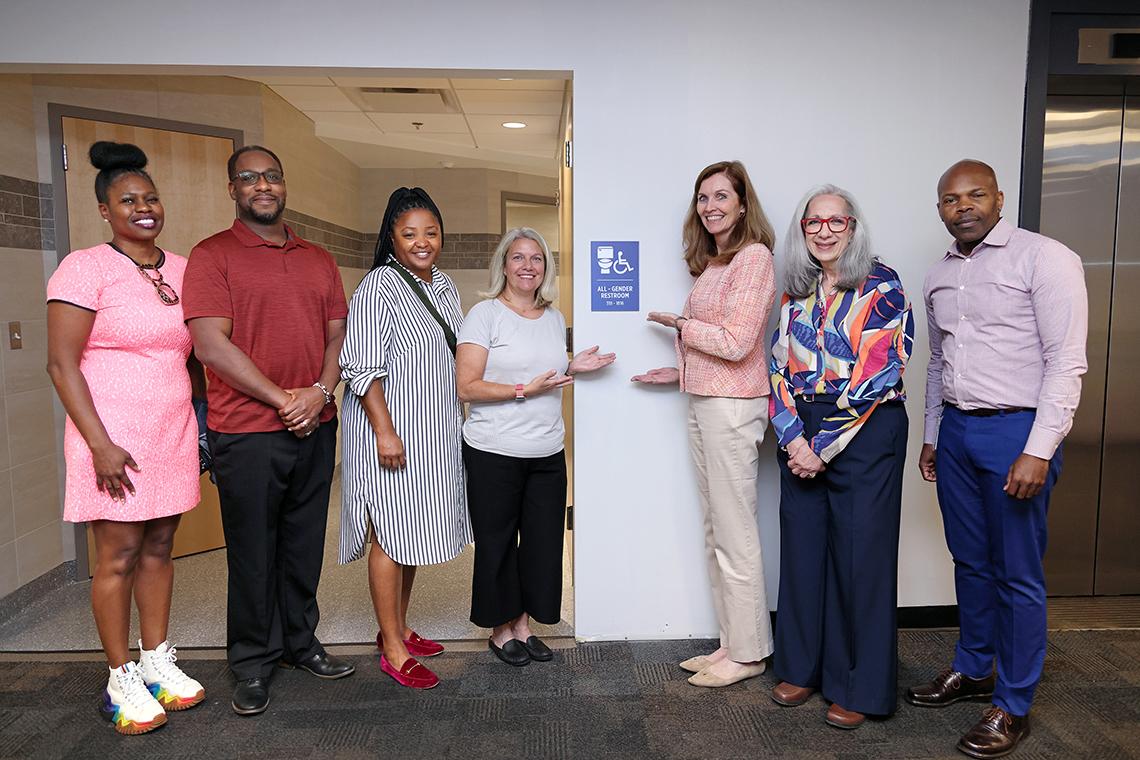 Group of people assembled in Bldg. 31 hallway pointing to "all-gender bathroom" sign on wall