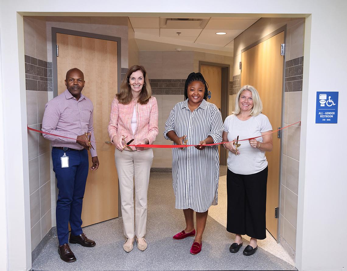 Group stands at bathroom entrance with large scissors to cut the ribbon.