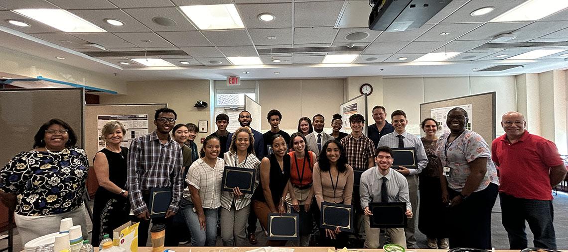 Students and their mentors pose for a photo in a large room. Some of the students hold dark-colored plaques.