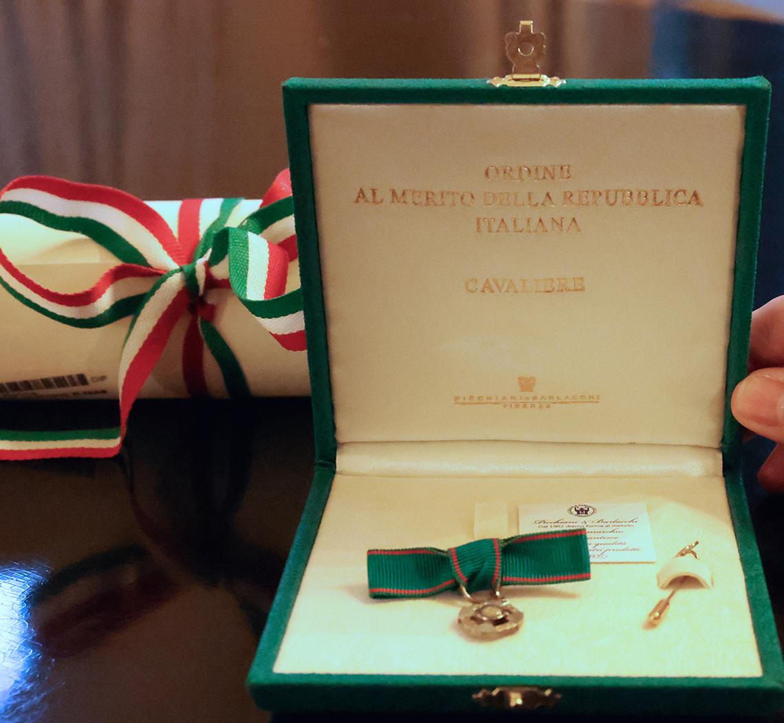 A medal displayed in an open box. A rolled-up certificate, tied with a red, white and green ribbon, sits behind the box.