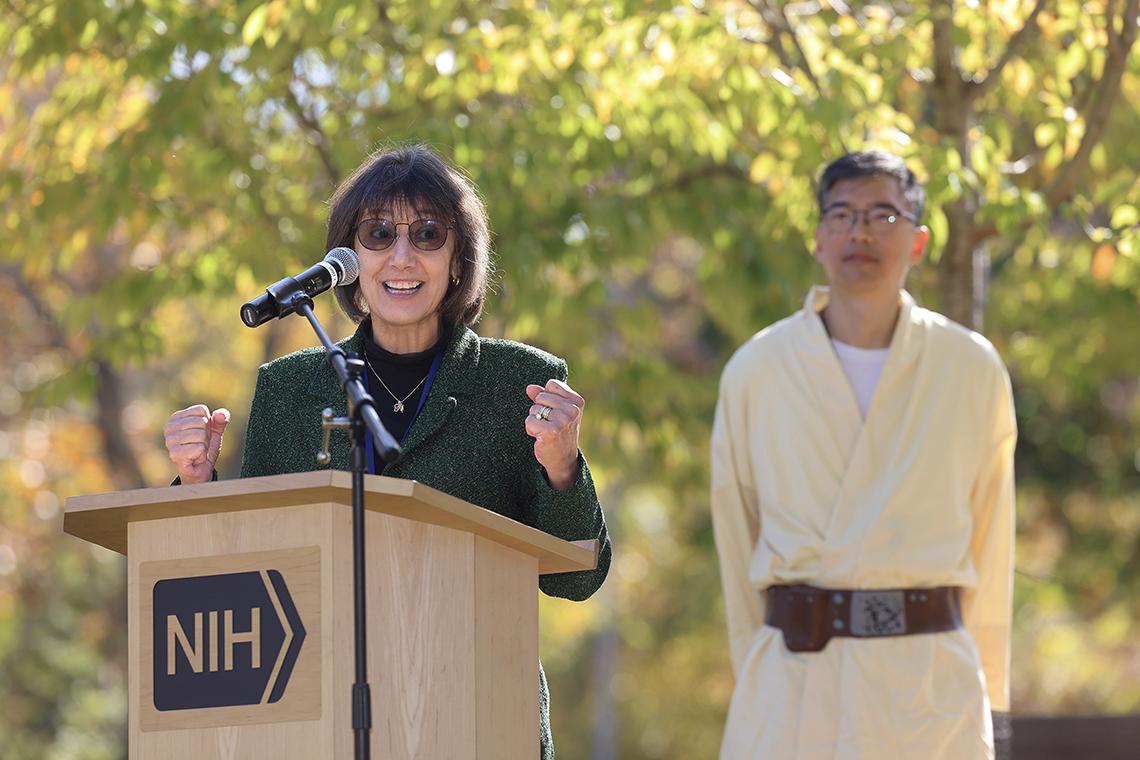 Bertagnolli speaks at a light-colored wooden lectern bearing the NIH logo while Chiang looks on.