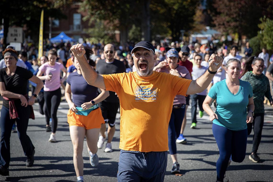 A runner in an orange t-shirt gestures enthusiastically; a large crowd of runners race behind and around him.