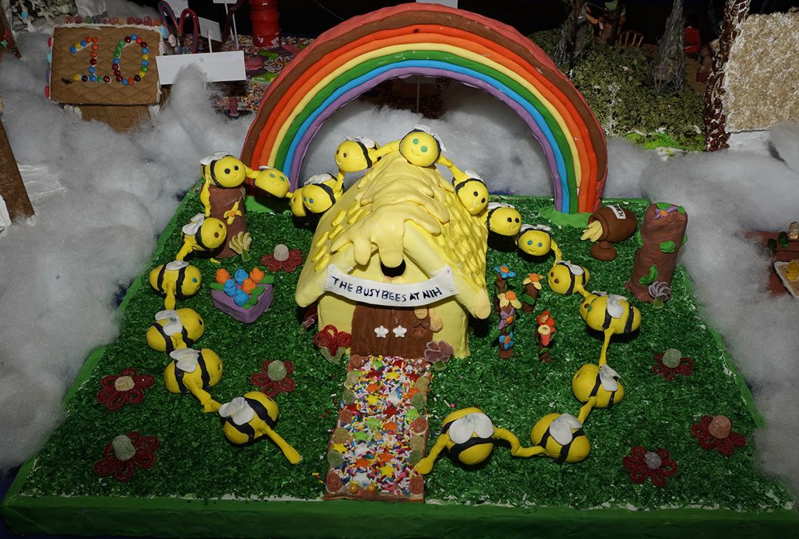 A gingerbread of bees flying around a small yellow house, titled "Busy Bees at NIH" with a rainbow arch