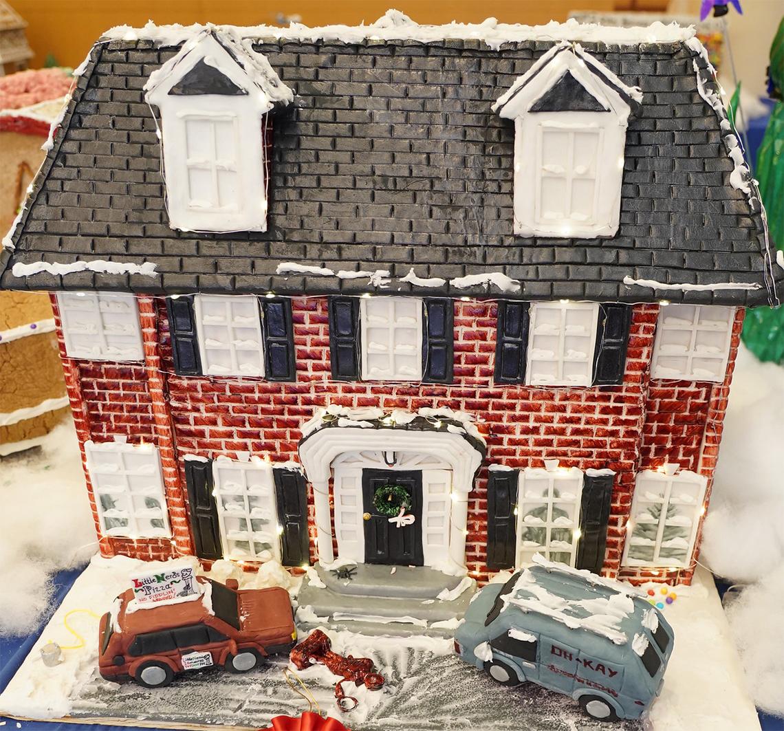 Gingerbread of a brick home with a pizza-delivery car, a van, and a man face down in the snow out front