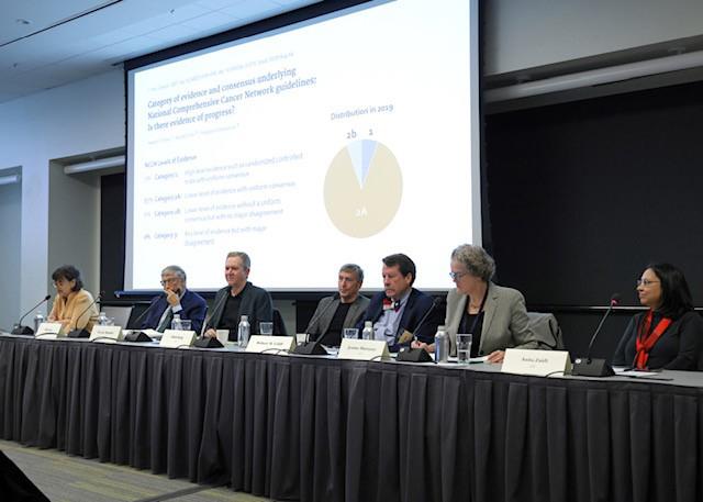 A line of seven speakers sit at a long table. A powerpoint presentation is visible behind them.