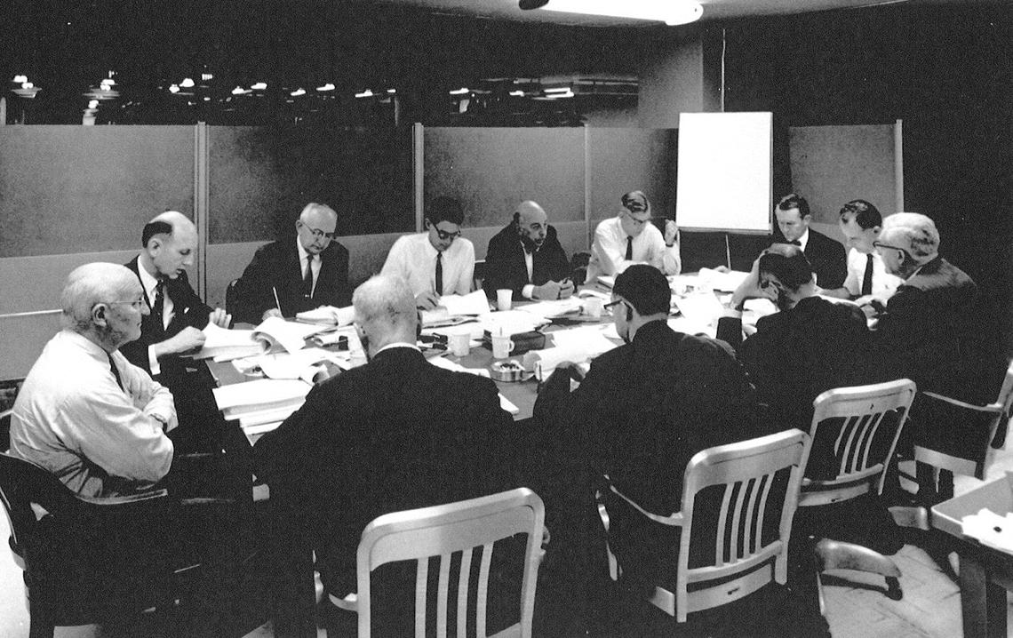 Black&amp;white image of 12 men seated around a conference table in a small enclosure, with wall open at top in background