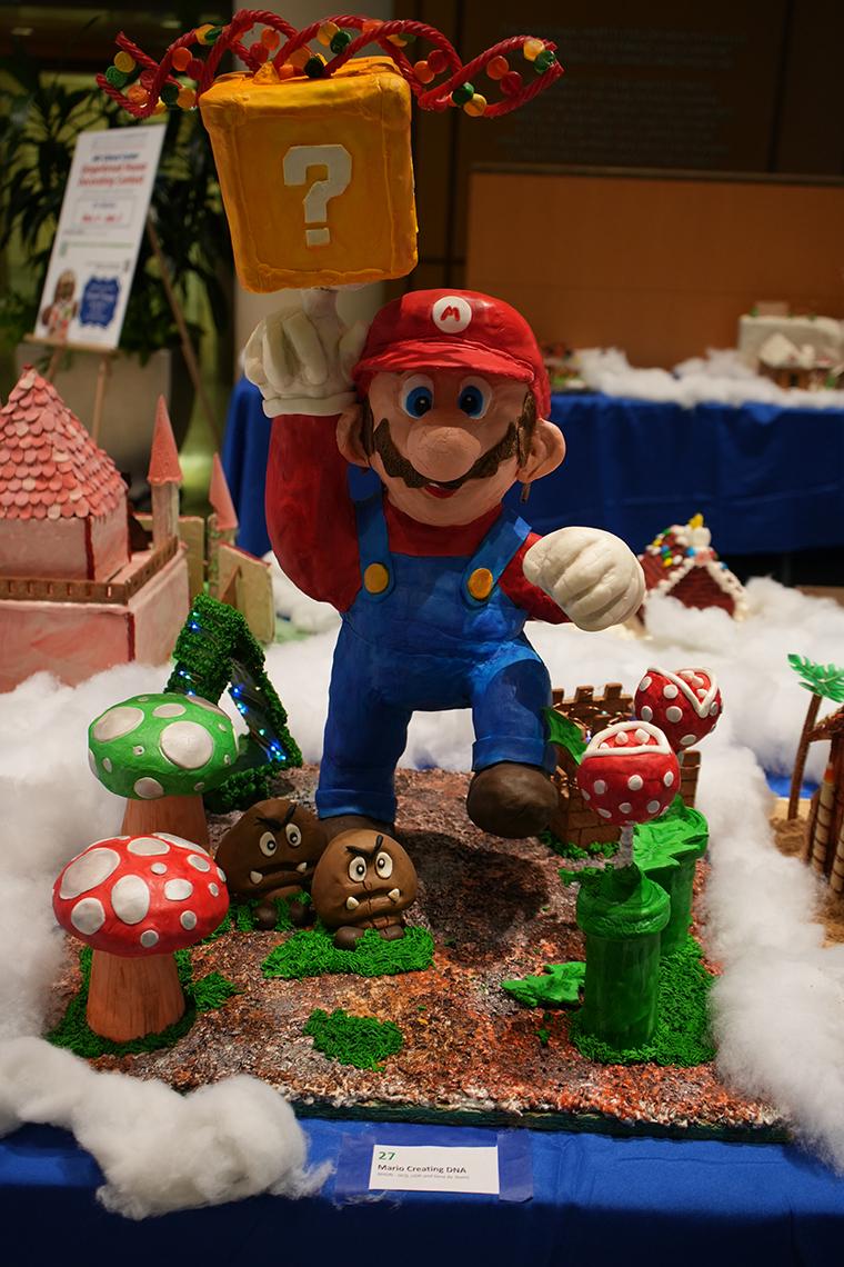 A large gingerbread of Super Mario, goombas, mushrooms and a castle with a candy double helix (DNA) above the yellow, question-mark block