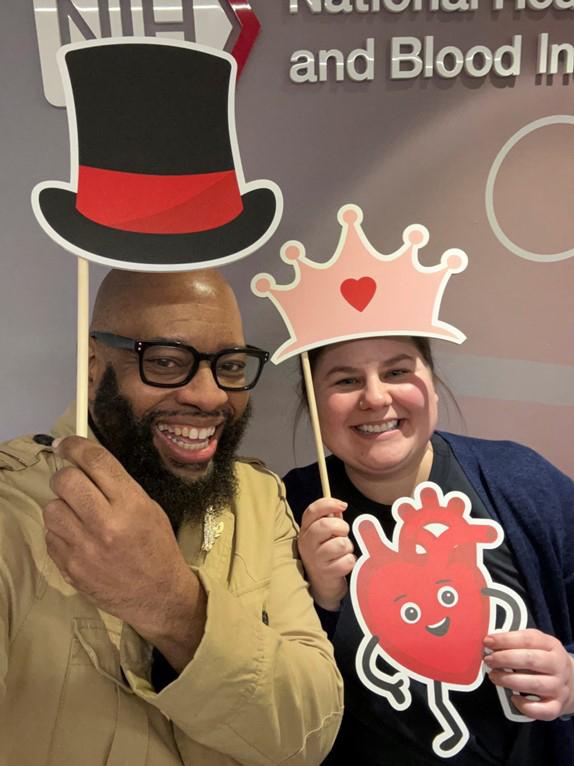Two people pose for a selfie with props and a cardboard cutout of a cartoon heart.