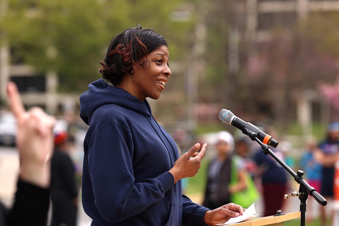 A woman in a dark blue hoodie gestures as she addresses a crowd.