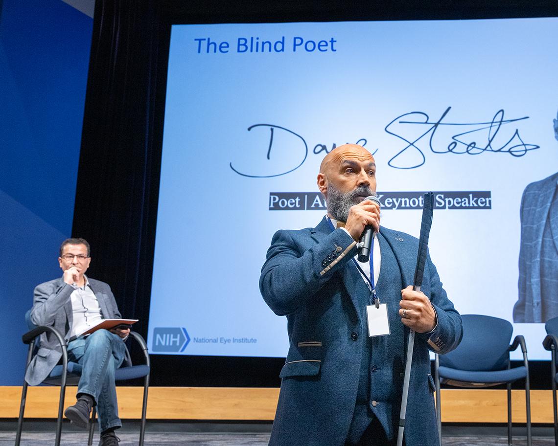 Steele speaks in an auditorium as Bharti looks on from behind