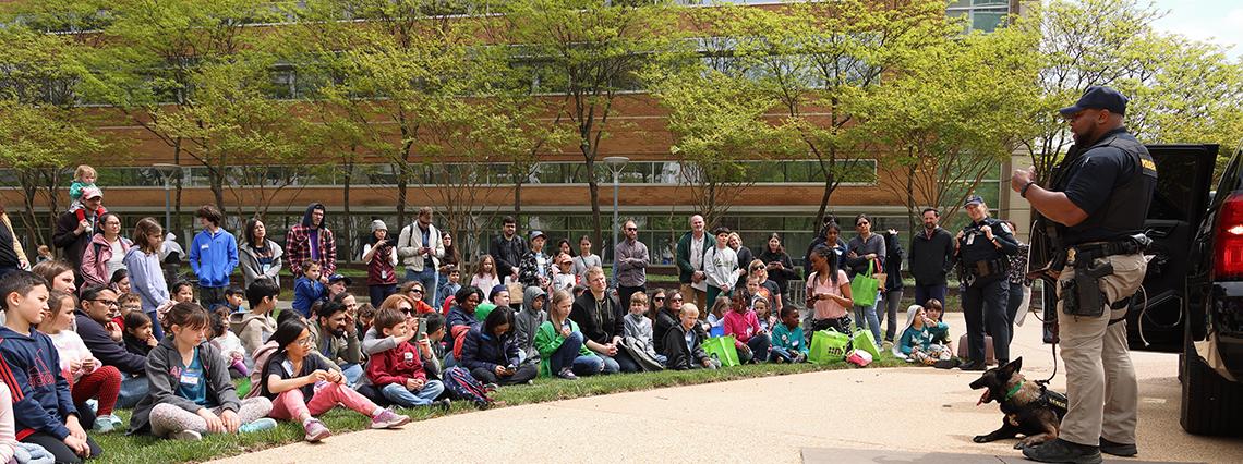 A large group of kids and parents assemble behind Bldg. 31 to watch canine demo with handler.