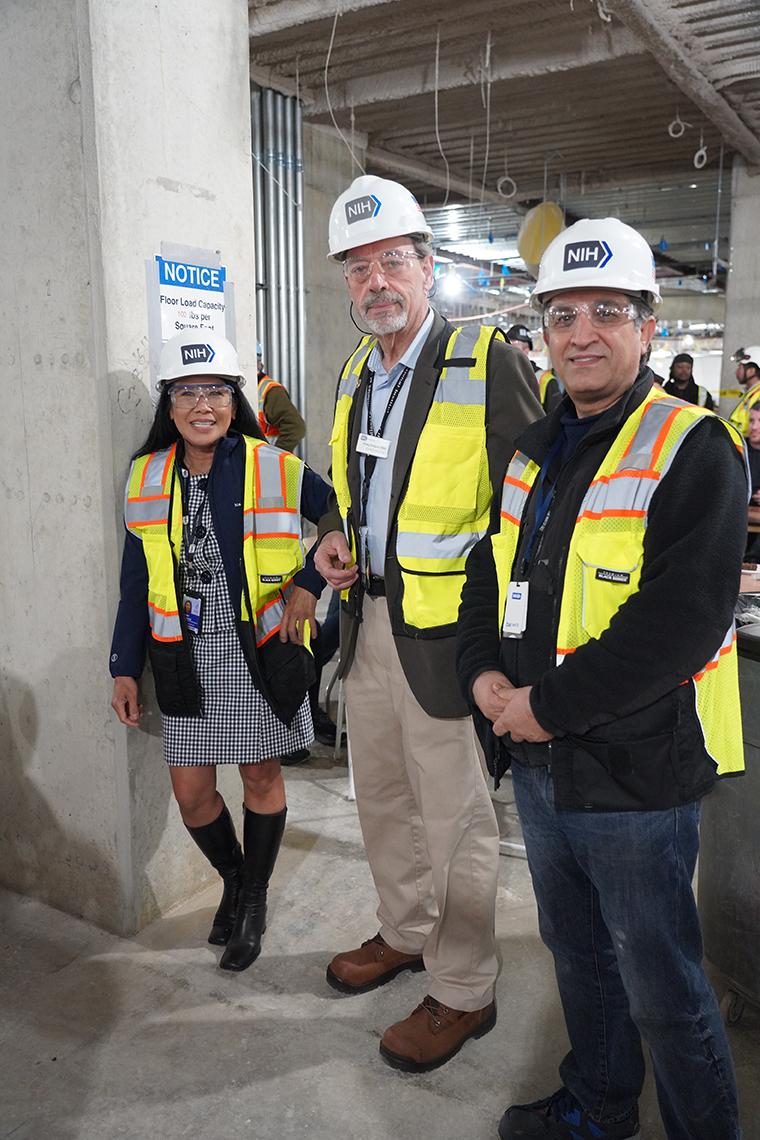 Three people in white hard hats and fluorescent yellow construction vests standing in the interior of a partly constructed building.