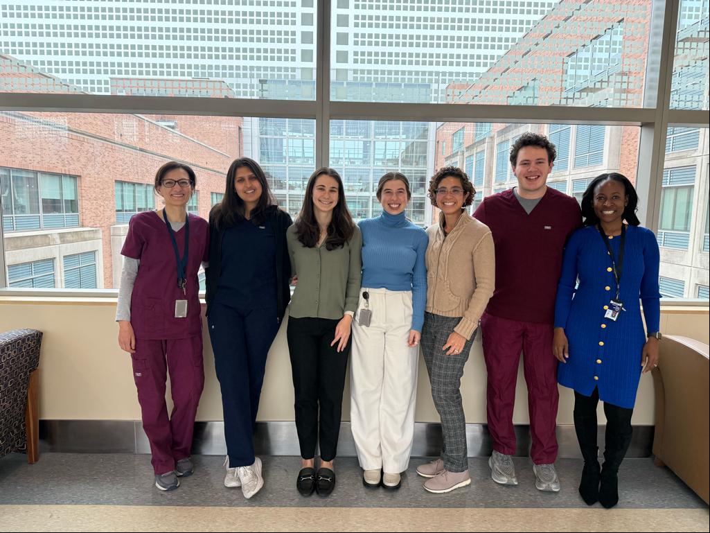 Seven people lined up against a window. Two wear burgundy scrubs and the rest wear office attire. A brick building is visible through the window behind them.