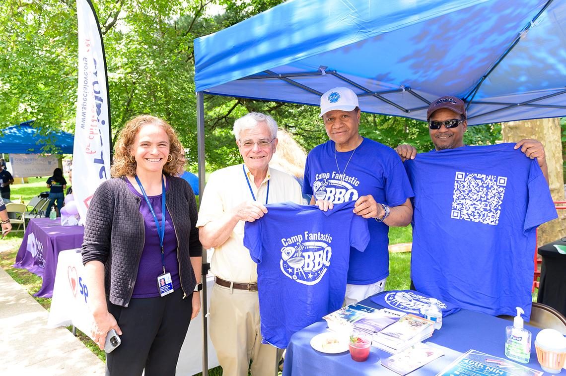 A man in the center holds up a t shirt. He is joined by his daughter to his left and a man to his right