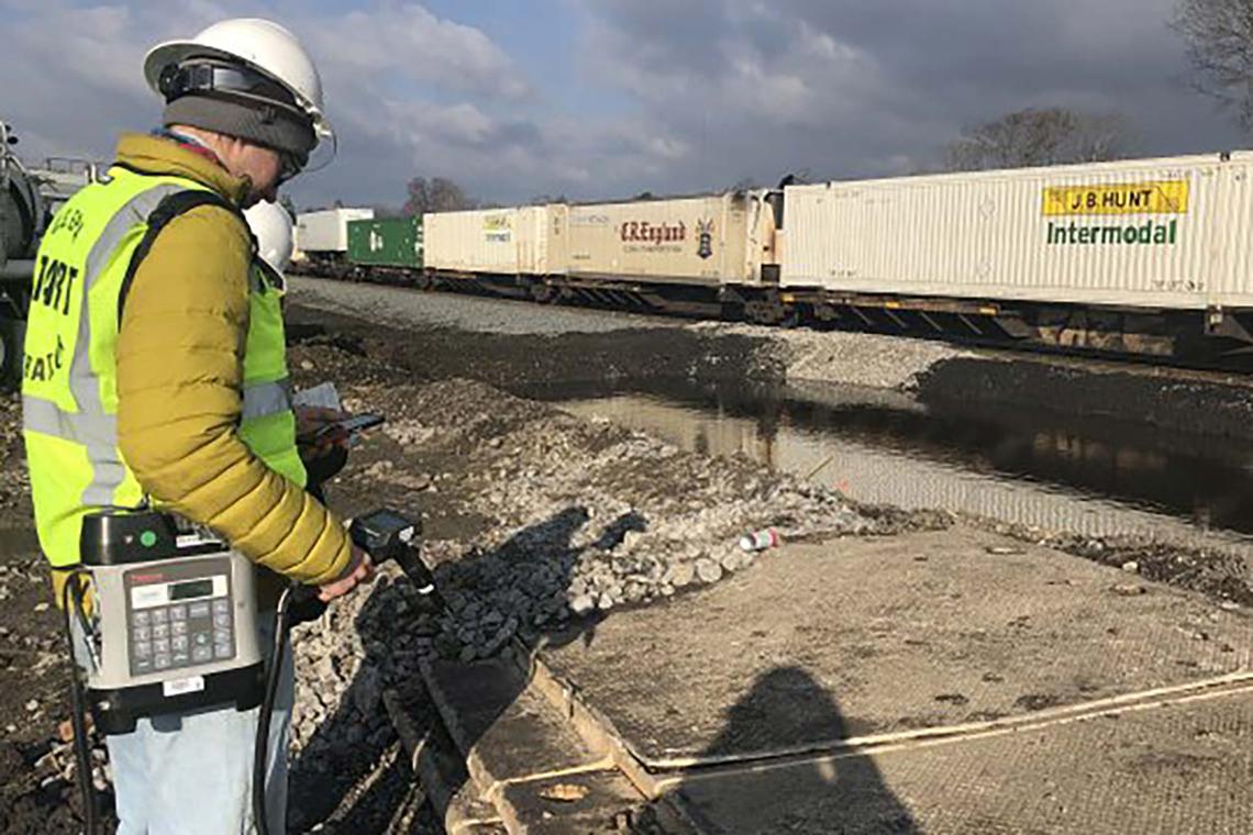 A person is a high-visibility vest stands near a passing train while monitoring air quality
