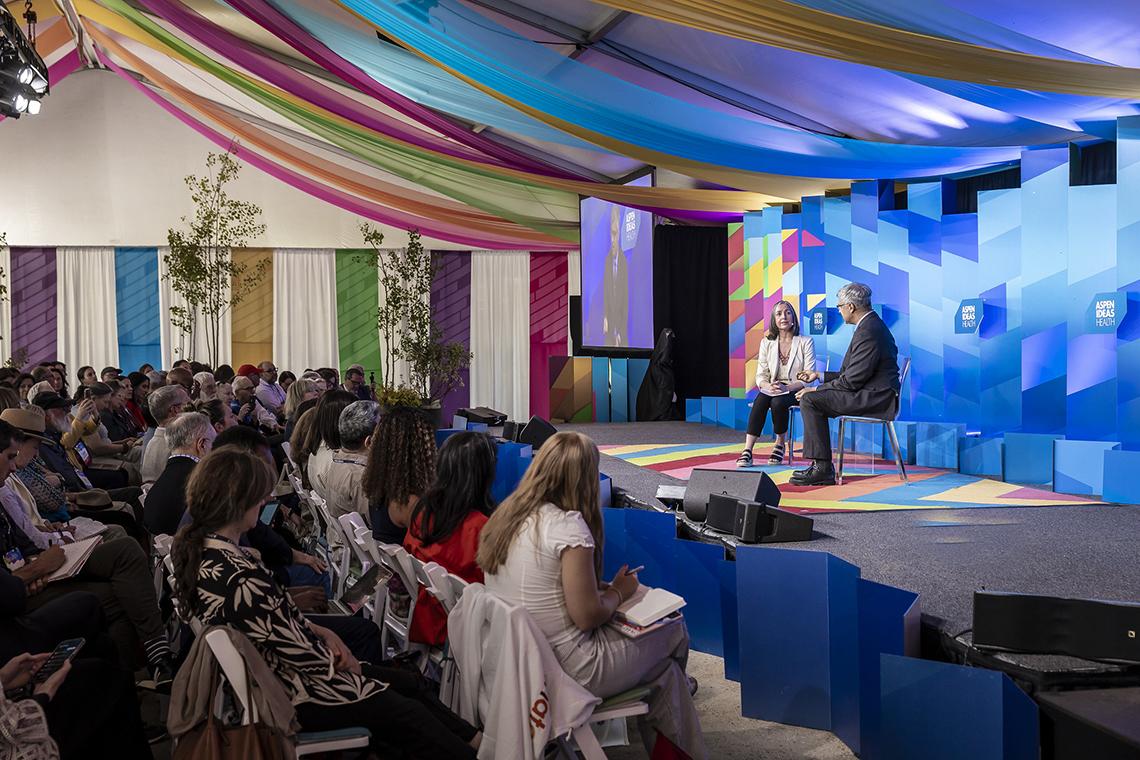 Reporter Ilgenfritz and Bhattacharya on stage in this wide shot showing audience and colorful fabrics hanging from ceiling