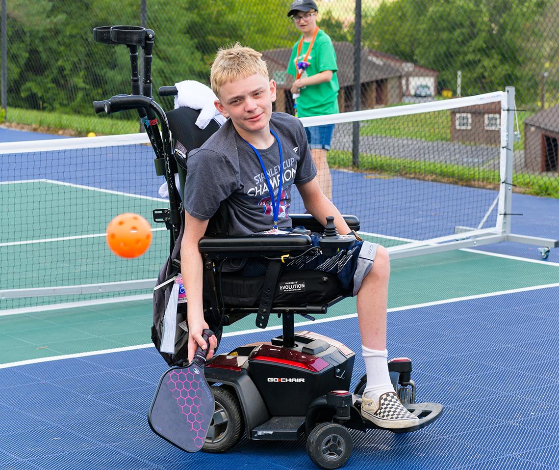 Child in wheelchair hits ball with racquet on pickleball court.