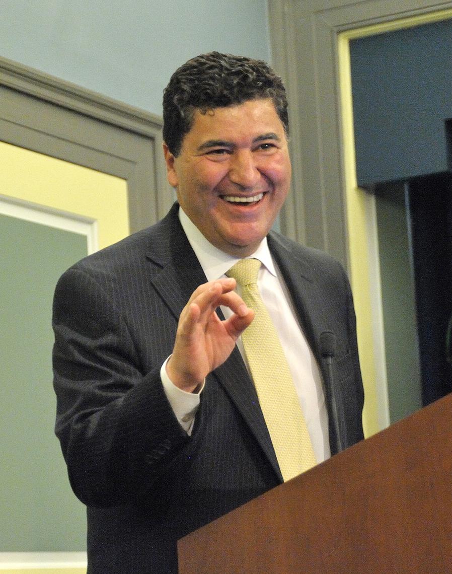 Zerhouni gestures as he speaks at a lectern. He is wearing a black suit jacket and yellow tie.