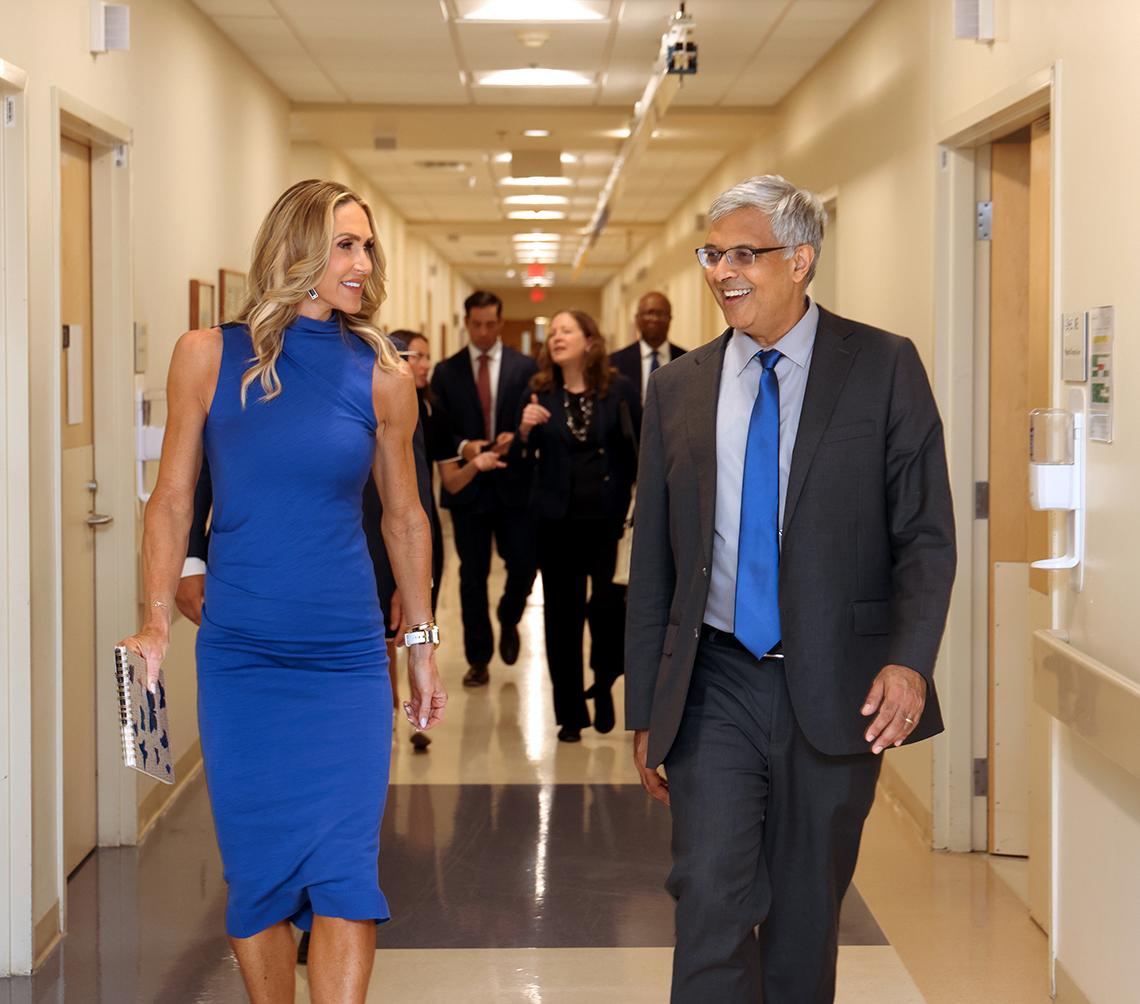Lara Trump and Jay Bhattacharya walk together, smiling, down a Clinical Center corridor.