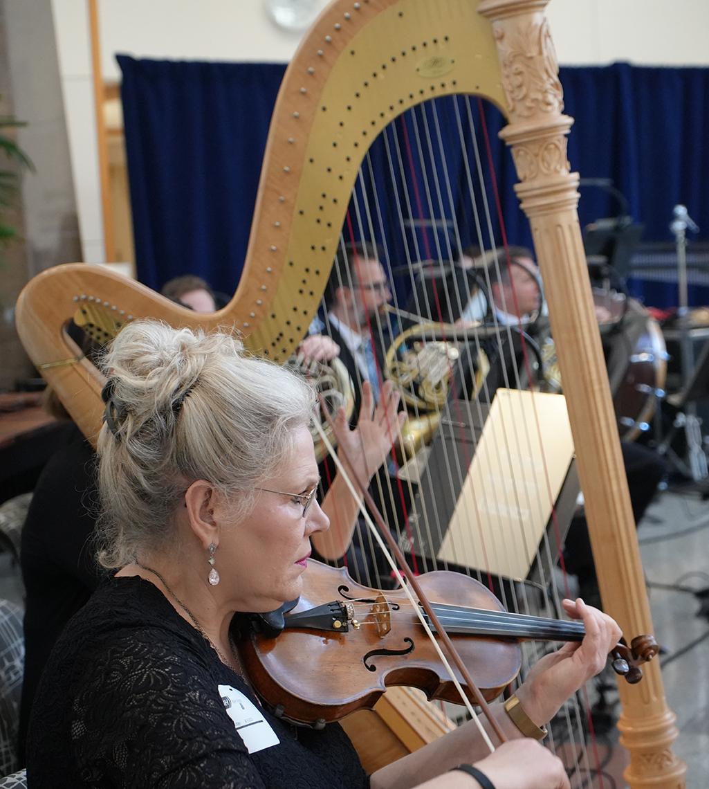 A violinist and harpist play as some horn players can be seen beyond the harp strings