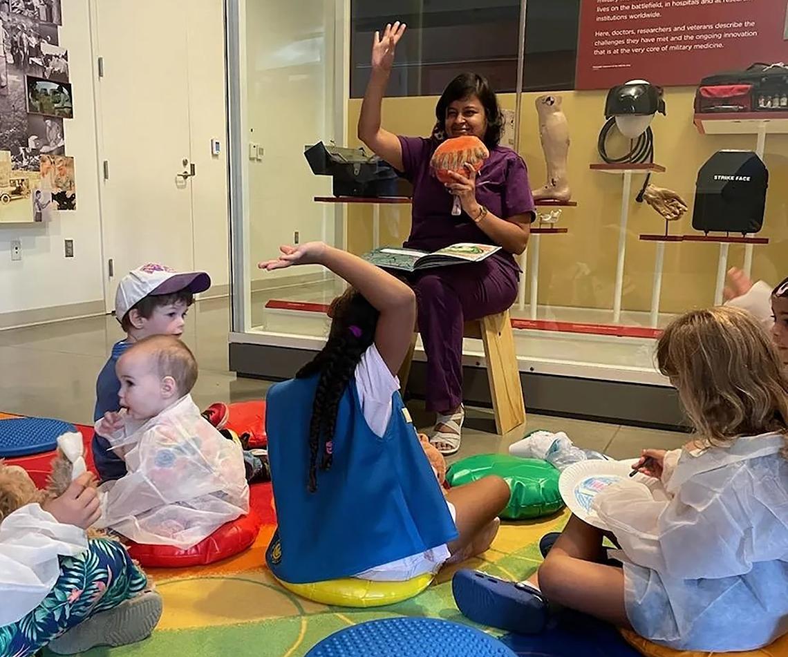 Viswanathan sits in a chair, holding a prop. Several children sit on colorful cushions at her feet.