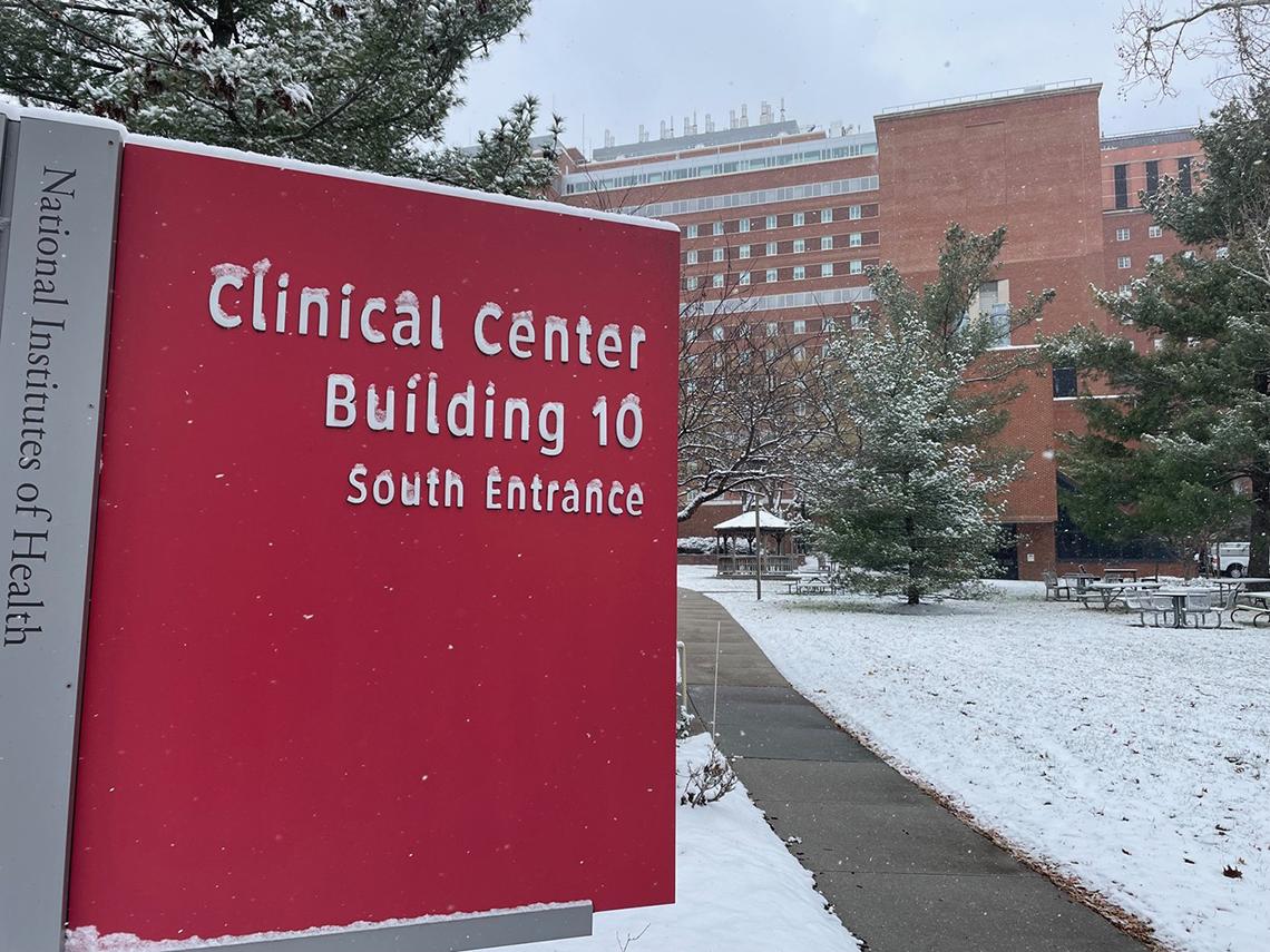A large red sign takes up the entire left foreground, reading "Clinical Center Building 10 South Entrance." A dusting of snow covers the top of the sign and the ground behind it. In the background is a multistory brick building.
