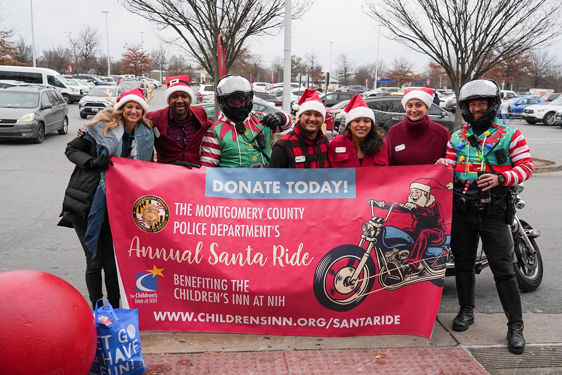 A group of people wearing Santa hats stand in a parking lot holding a red banner.