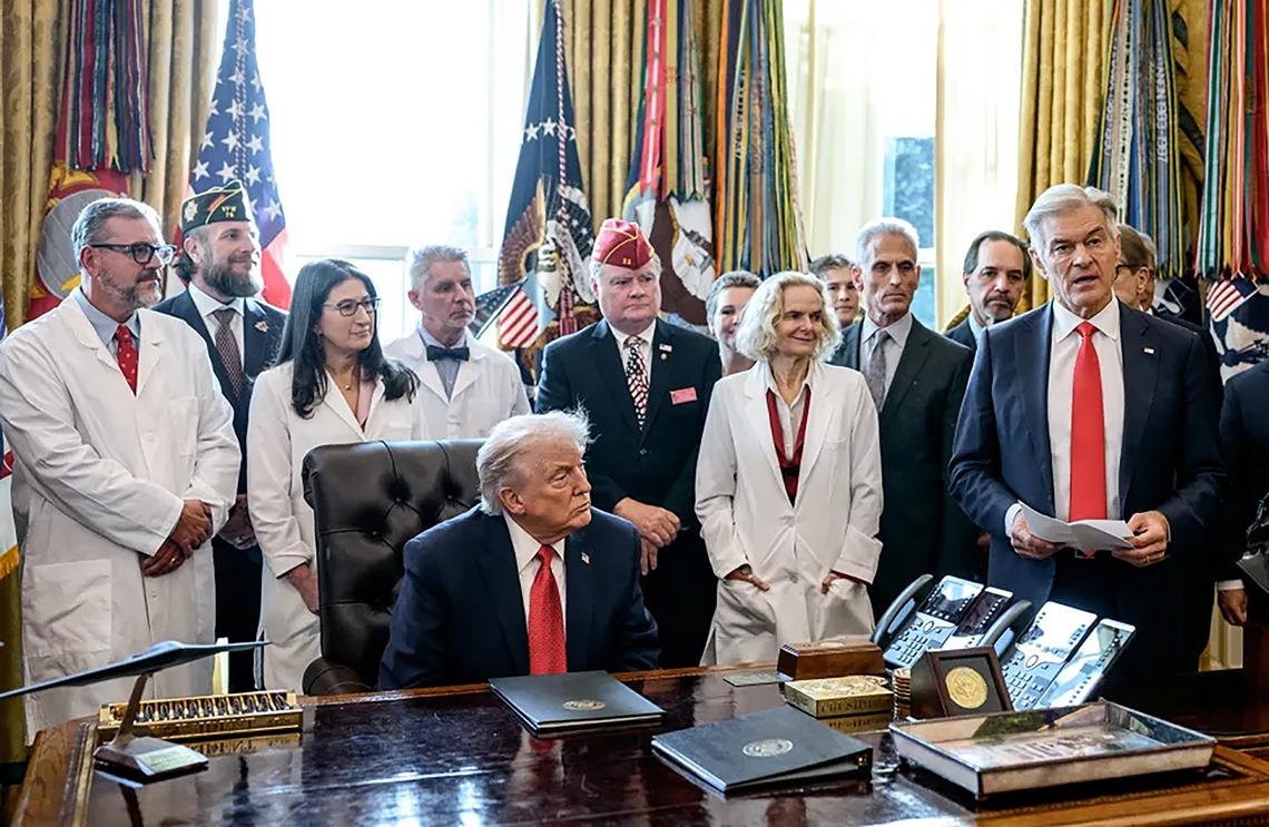 President Donald Trump sits at the desk in the Oval Office. A group of people, including Dr. Volkow, are gathered around him.