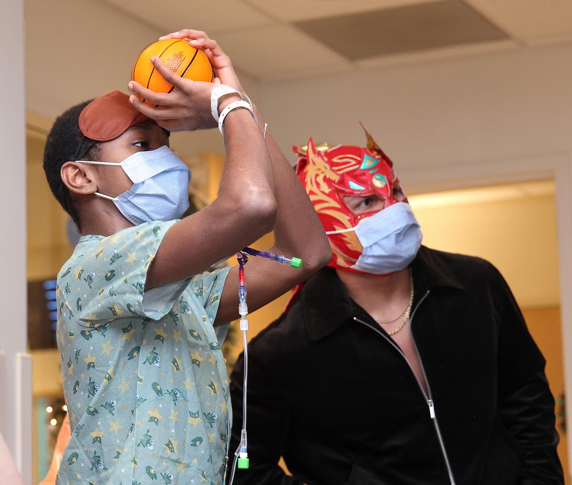 Child wearing mask, with IV cord dangling, throws a toy basketball as Lee looks on.
