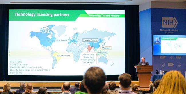 Wide shot of Whitehead at podium with audience viewing slide of world map showing vaccine technology transfer