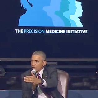 With mic in one hand, Obama gestures with other hand while seated in front of a projected slide.