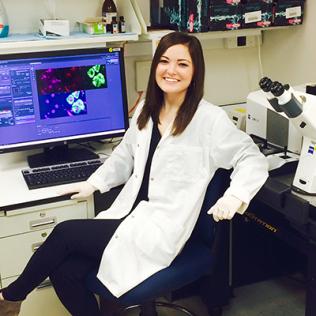 Stahl, in white lab coat, sits in front of computer monitor, next to microscope.