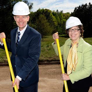 Wheeland and Birnbaum, wearing white hard hats, smile, holding shovels.