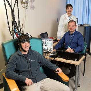 A young man sits smiling with a helmet of wires, as two doctors look on.