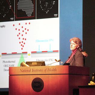 West at the NIH podium with slide projected beside her